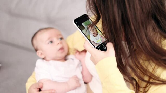 Young mother hold her little baby infant and taking photograph saving great memories on smartphone mobile phone at light home Happy motherhood concept