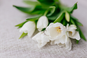 Obraz na płótnie Canvas An engagement ring in white gold with a diamond lies in a bouquet of white tulips. Gift for Women's Day, Valentine's Day. Beautiful spring background with flowers