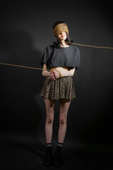 Portrait of a poor skinny girl in black crumpled clothes with a rope wrapped around her head and hands, which is pulling her somewhere. concept of poverty, helplessness, oppression, or captivity.
