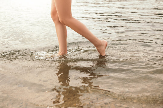 Close up naked slim woman legs walking barefoot along the river. Female feet on sea beach with splashing waves and foam. Girl tourist on vacation. Lady go on shore after bathing

