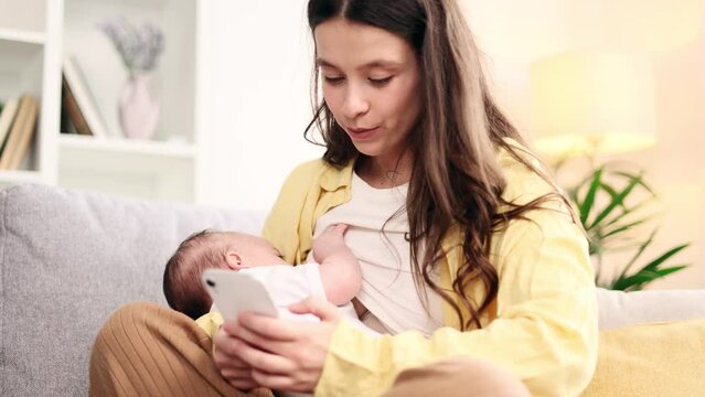 Attractive young woman mother breastfeeding her baby while using smartphone mobile phone scrolling social media chatting texting browsing online or using motherhood app application at light home