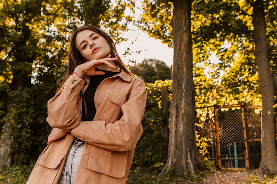 Caucasian Young Pretty Stylish Woman Standing Outside Street Dressed Brown Trench Coat Smiling Poses Outside City, Spring Autumn Season Nature Sunshine Light Leaks. Cute Brunette Hair Lady 
