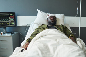 African American soldier with his head wrapped by bandage lying in bed between dropper and computer...