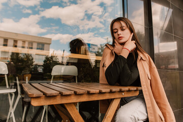 Fototapeta premium Caucasian young pretty stylish woman sitting table near street cafe restaurant chill dressed brown trench coat smiling happy poses outside in city, spring autumn season. Cute brunette hair lady 