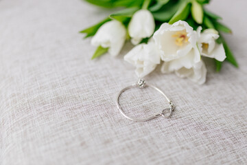 Bracelet in white gold lies inext to a bouquet of white tulips on white background. Gift for Women's Day, Valentine's Day. Beautiful spring background with green leaves. Mock up