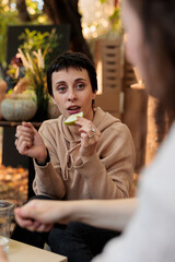 Positive woman enjoying organic fruits and veggies taste at local street food market, sunny fall day. Female farmer sitting at table with consumer, offering sample of natural homegrown produce.