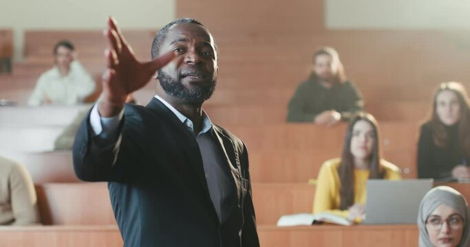 African American male professor standing in front of students and giving lection. Man teacher explaining lesson subject in auditorium. Close up. Teaching work. Working at University. Explanation.
