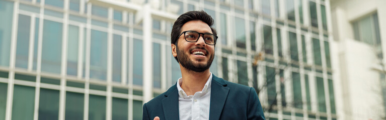 Smiling indian businessman in suit and glasses with laptop near office building