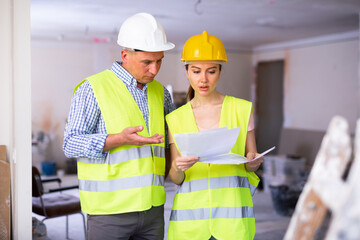 Man and woman construction workers in protection gear holding documents ant talking on indoor building construction site