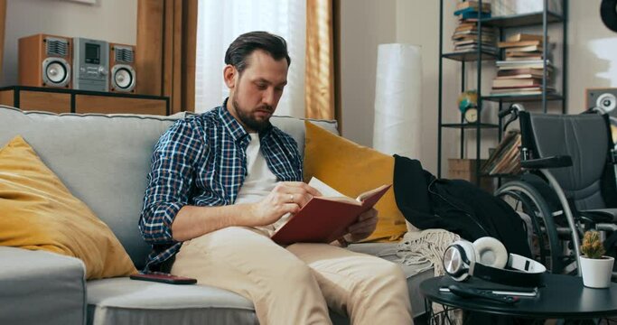 Young handsome man sitting on the sofa reading book spending free time alone at home disabled person with wheelchair feeling upset sad.
