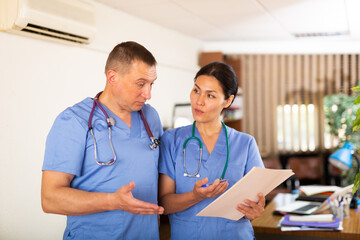 Two professional doctors standing in office, reading medical card of patient and discussing diagnosis