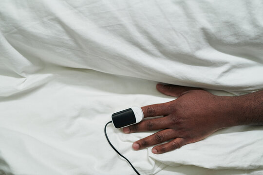 Hand of young African American male patient with pulse oximeter on fingertip lying under white cotton sheet in bed in hospital ward
