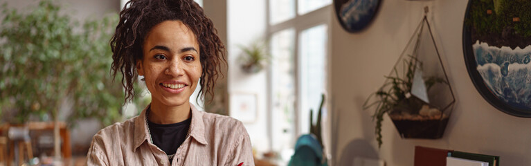Attractive smiling florist stands with crossed arms on background of her own floral decor studio