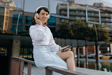 Brunette woman in white shirt, blue skirt is holding phone outside. Stylish woman in headphones listening to music on city buildings backdrop