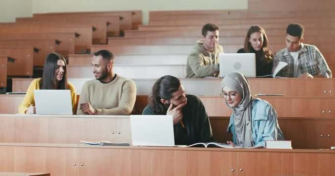 Mixed-races males and females students sitting in auditorium with laptops, talking and working on task at college. Multiethnic guys and girls at computers discussing studying project at high school.