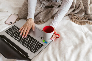 A beautiful girl sitting on a bed wrapped in a blanket with a warm drink in a mug and working on a...
