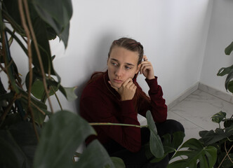 A teenage girl is sitting in the corner by the wall in the room next to the houseplants. She's upset and sad. The problem of loneliness, bullying, mental health in the modern world.