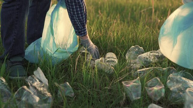 Group of people are cleaning up plastic garbage in park on grass. Hand of volunteer picks up plastic bottle from grass in meadow and puts it in garbage bag. Human pollution of nature. Friendly planet