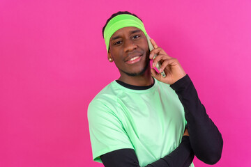 Fototapeta premium Black ethnic man with a phone in green clothes on a pink background, smiling talking on the phone