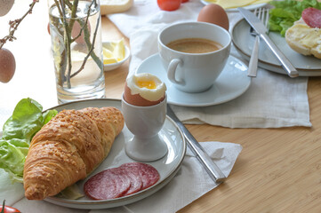 Breakfast or brunch with croissant, cooked egg, sausage, cheese and coffee on light napkins and a wooden table, selected focus