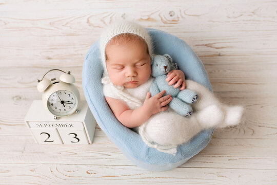 Top View Of A Newborn Baby Girl Sleeping In A White Jumpsuit With A Blue Teddy Bear, In A Blue Felt Chair. With An Autistic White Alarm Clock. Beautiful Portrait Of A Little Girl 7 Days, One Week.