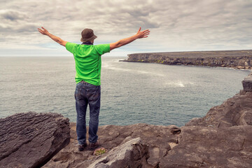 Male tourist in green shirt standing on edge of a cliff hands up in the air, looking at stunning scenery. Aran Island, Ireland. Travel and tourism. Rough Irish nature landscape.