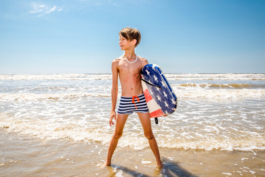 Young boy in striped swim trunks standing in the water at the beach with his boogie board