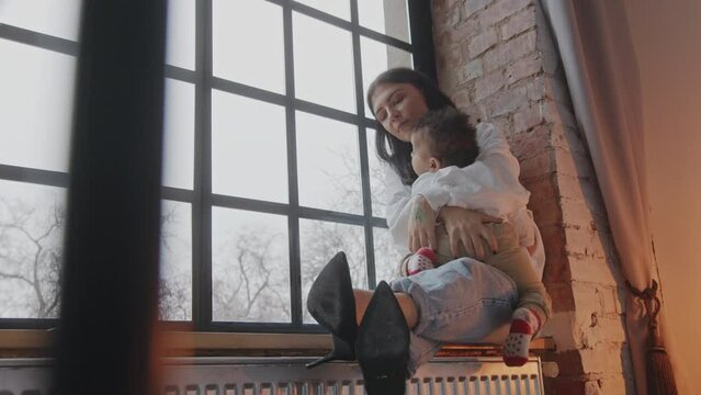 young Caucasian mother and her cute little son sitting on the window sill, hugging each other and enjoying. High quality 4k footage