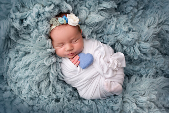 Top view of a newborn baby girl sleeping in a white children's winding with flowers on a blue carpet flakati background and a blue heart. Portrait of a little girl 7 days, one week
