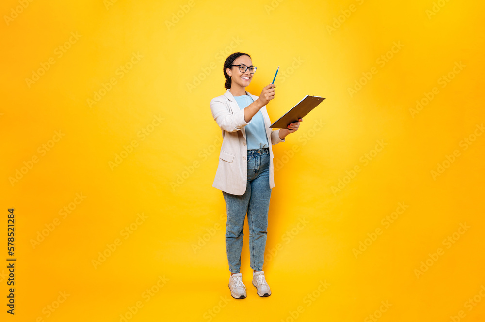 Wall mural Full length photo of pretty young latino or brazilian woman, in stylish casual wear, hr manager, real estate agent, holds folder with papers document, looks away and smile, isolated orange background