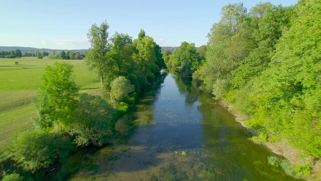 AERIAL: Slowly flowing river surrounded with trees meandering across the field. Stunning water reflections of blue sky and lush greenery on the river banks. Picturesque location for summer adventures.