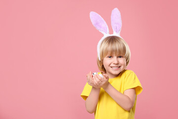 Obraz na płótnie Canvas Happy boy in bunny ears headband holding painted Easter eggs on pink background. Space for text