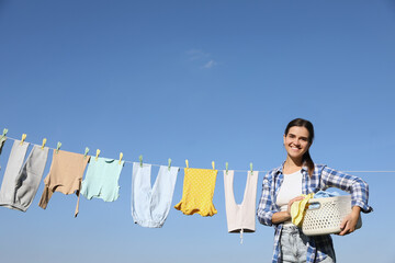 Smiling woman holding basket with baby clothes near washing line for drying against blue sky...