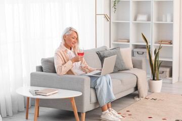 Mature woman with glass of wine using laptop on grey sofa at home