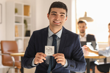 Young man holding sticky paper with text HAPPY FOOL'S DAY in office