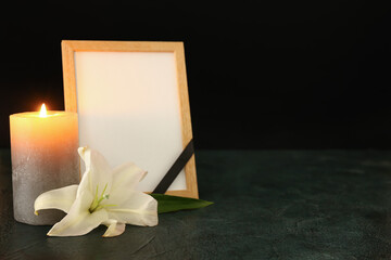 Blank funeral frame, burning candle and lily flower on dark table