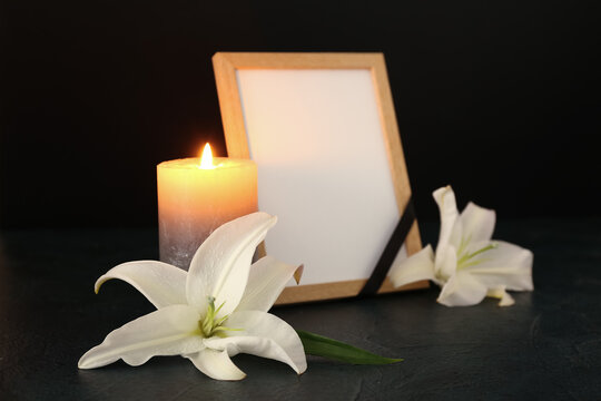 Blank funeral frame, burning candle and lily flowers on dark table