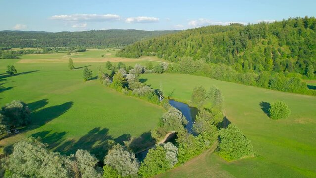 AERIAL: Charming Planina Karst Field with meandering river Unica on a summer day. Lush forested hills surround beautiful valley with vibrant green grasslands and lush greenery on winding river banks.