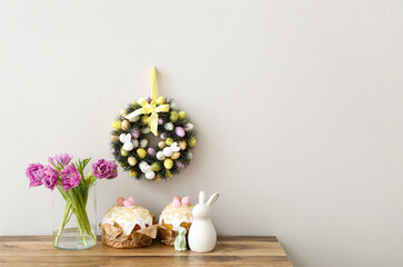 Table with tulips in vase, Easter cakes, rabbits and wreath on light wall