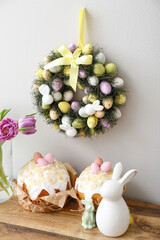 Table with tulips in vase, Easter cakes, rabbits and wreath on light wall