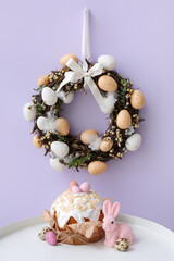 Table with Easter cake, rabbit and wreath on lilac wall