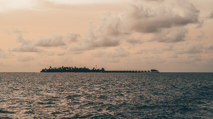 Shot from a boat sailing in the sea, this low-key sunset offers a mesmerizing view of an island in the Maldives. The silhouettes of overwater bungalows in the far back enhance the beauty of the scene