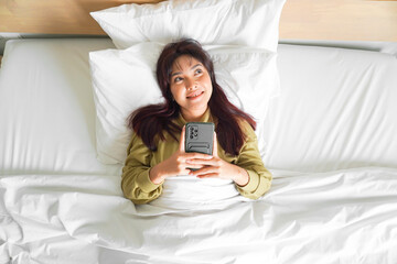 A thoughtful young Asian woman is holding her phone while lying on the bed