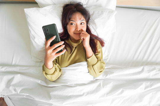 A thoughtful young Asian woman is holding her phone while lying on the bed
