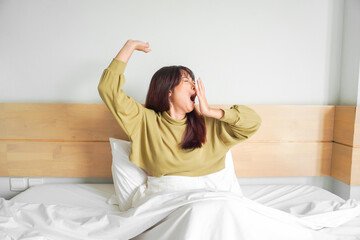 Cute Asian woman stretching out and yawning while sitting on her bed in bright bedroom