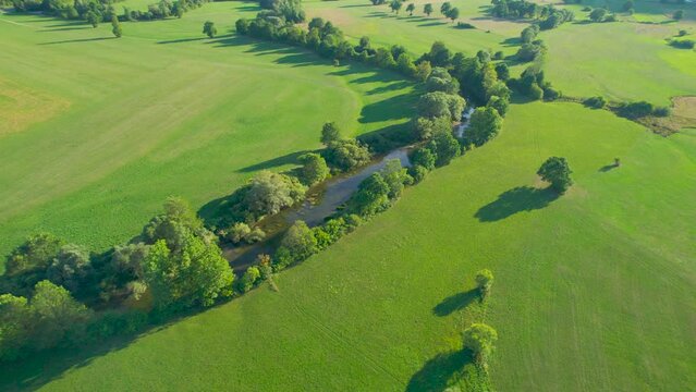 AERIAL: Meandering river, surrounded by lush greenery, winds through the fields. Picturesque countryside with slow-flowing river and vibrant green meadows in flattering summer afternoon sunlight.
