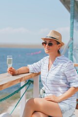 A woman in a straw hat and sunglasses sits in a restaurant with a glass of champagne overlooking the sea.