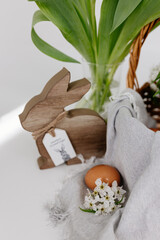A wicker basket, on a gray linen napkin, are painted eggs and white branches with blossoming pear flowers, an Easter wooden rabbit an glass vase with white fresh tulips. Spring Easter background