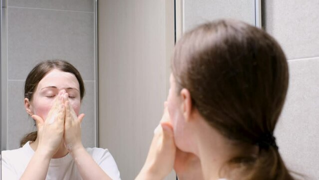 Young woman stands in front of a mirror in the bathroom and applies a facial wash on her face close-up.