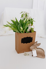 The Wooden Easter Bunny sits near a gift bag with white tulips on a light white background. Spring Background for Easter. International Women's Day, March 8th